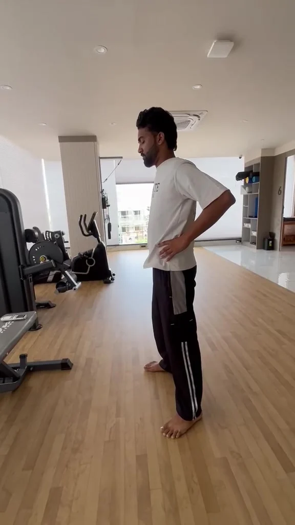 Man stands upright in a gym, head bowed, both hands resting on his lower back
