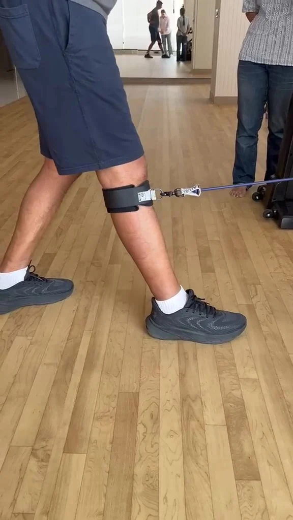 Leg with a cuff around the upper calf below the knee and a stretched cord mid-step, with other people in the background.