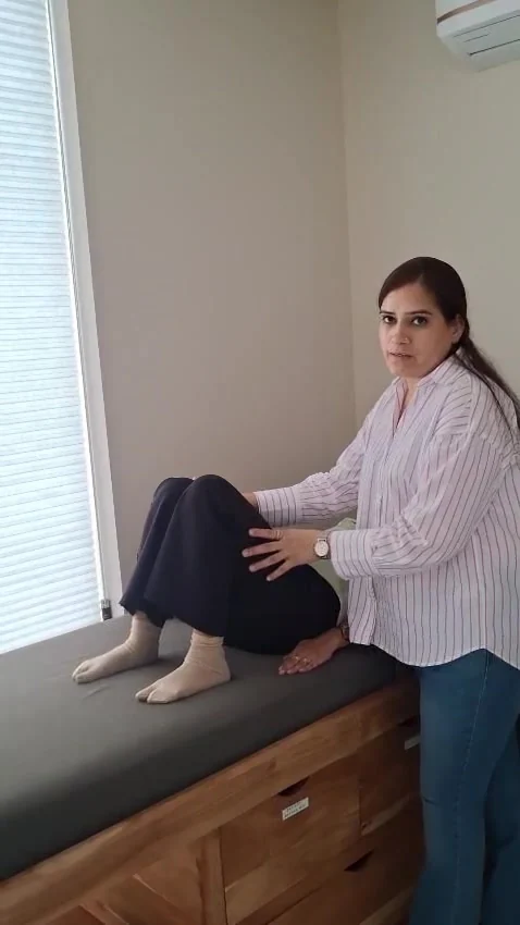 Woman looks at the camera with one hand resting on the bent knee of a person sitting on a treatment couch with both feet flat on the surface