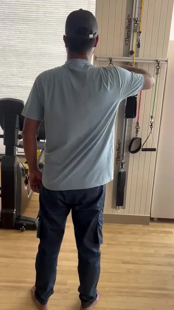 Man photographed from behind with one arm raised to a wall-mounted resistance band rail in a clinic gym.
