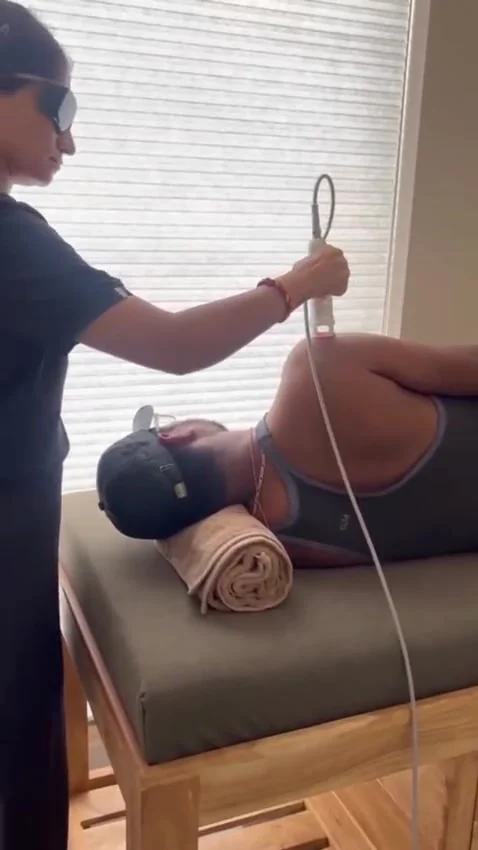 Patient on their side with a cap and eye shield covering the upper face while a therapist holds a laser probe over the shoulder