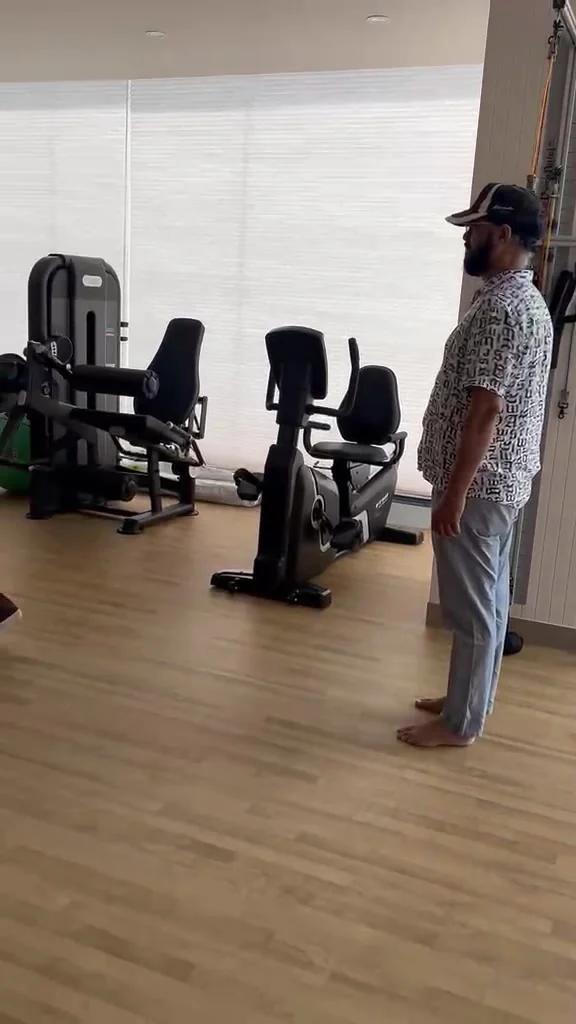 Bearded man in a cap and printed shirt standing barefoot in profile on the wooden gym floor, with a black weight-stack leg machine and a recumbent exercise bike across the room in front of him.