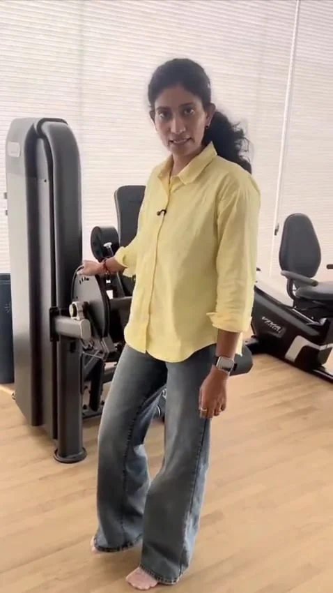 Woman in yellow shirt stands barefoot beside a gym weight machine, hand on its frame