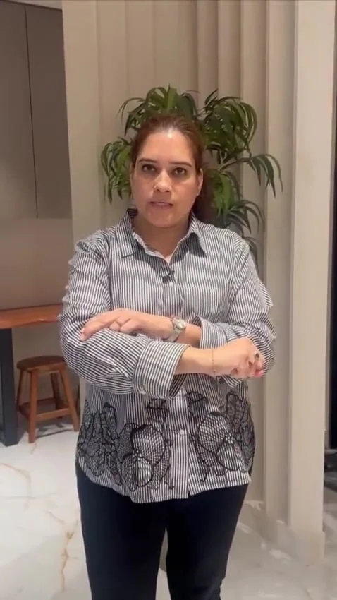 Woman in a striped shirt stands indoors with her arms folded, one hand resting on the opposite forearm, speaking to the camera